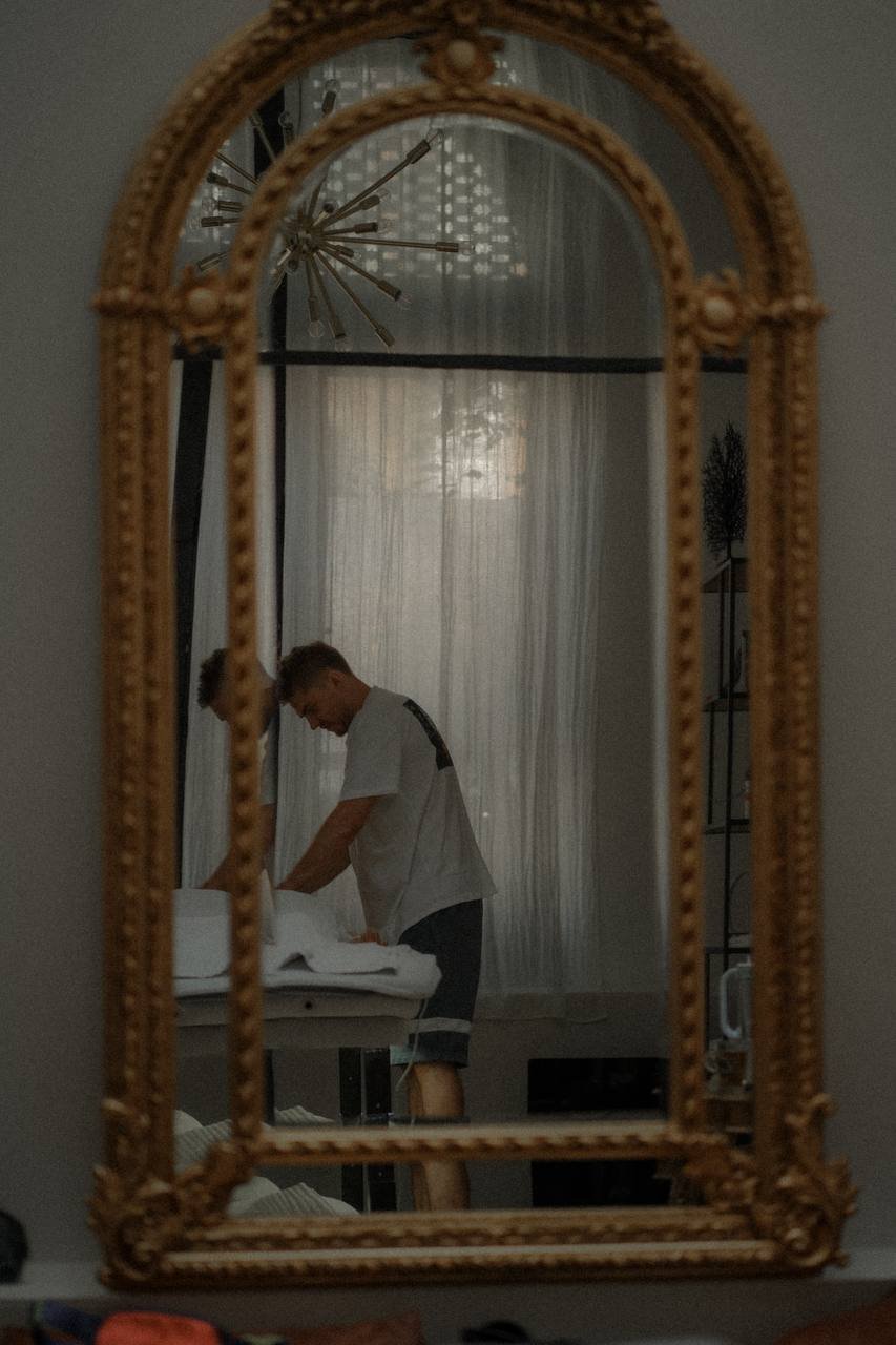 A person is arranging or making a bed in a room, viewed through an ornate mirror frame.